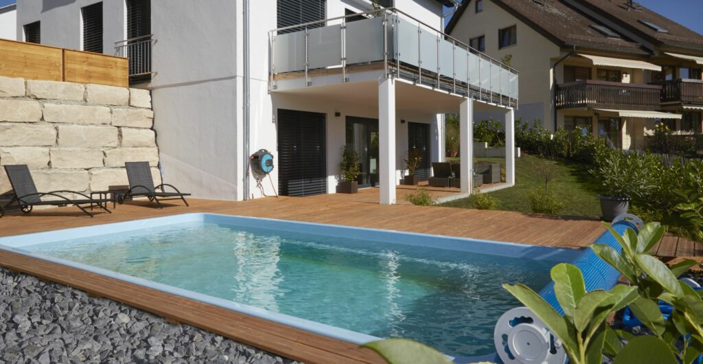 Ein modernes Haus mit Balkon überblickt einen rechteckigen Außenpool, der von einer Holzterrasse umgeben ist. Am Pool stehen zwei Liegestühle, und im Hintergrund ist ein weiteres Haus zu sehen, das eine beeindruckende Gartengestaltung zeigt.
