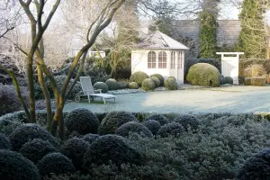 Frostbedeckter Garten mit gestutzten Büschen, einem weißen Pavillon und einem einzelnen Stuhl.